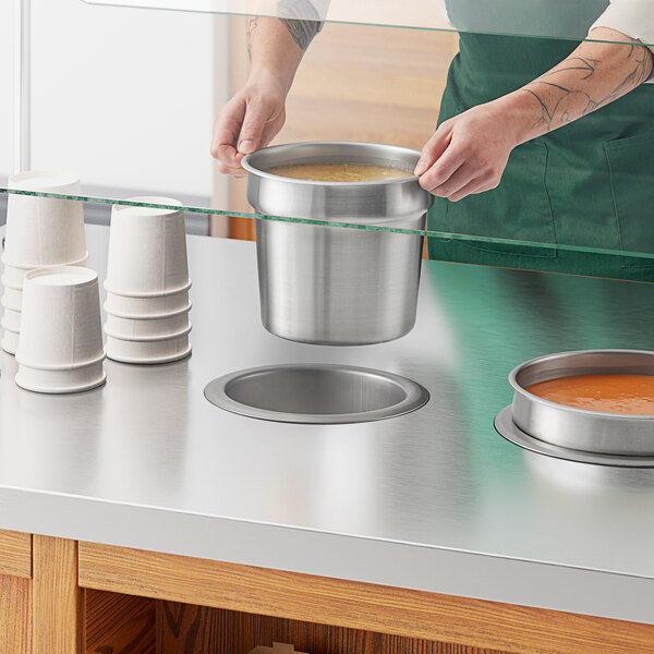 A person holding a silver pot of soup over a white Avantco drop-in well on a counter.