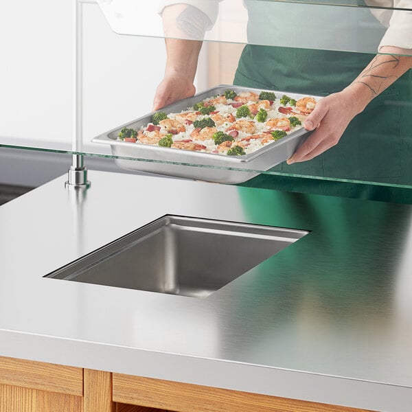 A person holding a tray of food with shrimp and rice over a metal drop-in hot food well.