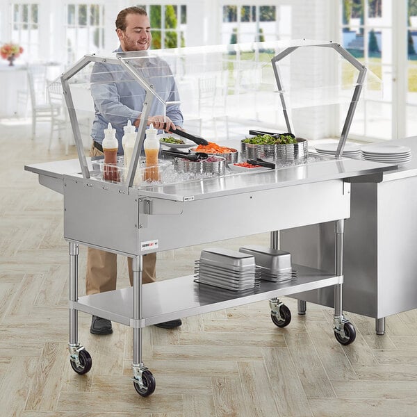 A man standing behind a stainless steel ServIt ice-cooled food table with a sneeze guard and side trays.