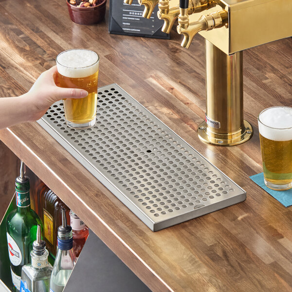 A rectangular stainless steel surface mount beer drip tray with circular holes, placed on a wooden bar counter next to a beer tap.