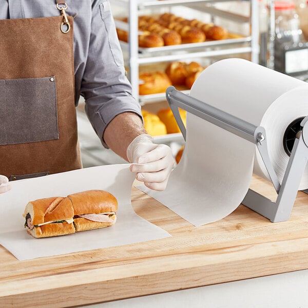 A person in a brown apron and gloves using Choice 55 lb. Premium Wet Wax Paper to wrap a ham and cheese sandwich.