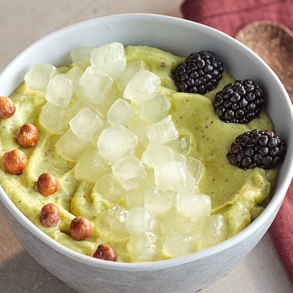 A bowl of green smoothie with blackberries and ice cubes, with Pitaya Foods IQF Aloe Vera Pieces on the table.