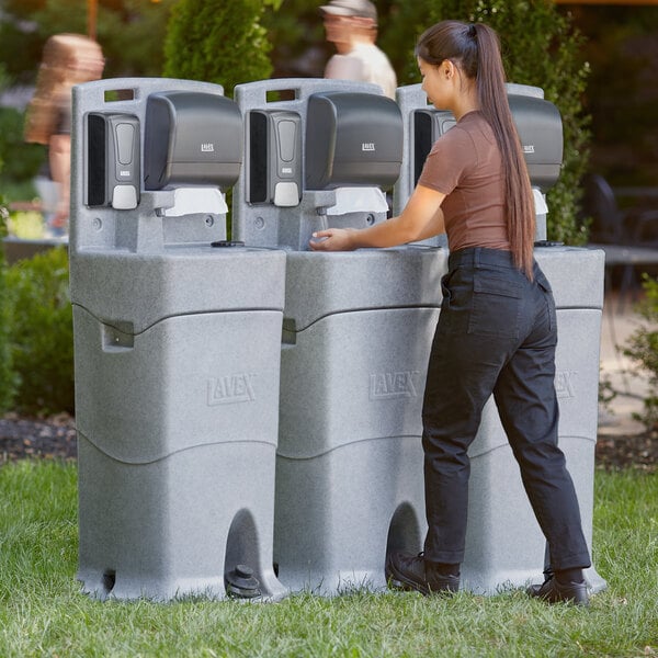 A woman using a Lavex mobile hand wash station with foam soap and paper towel dispensers in an outdoor catering setup.