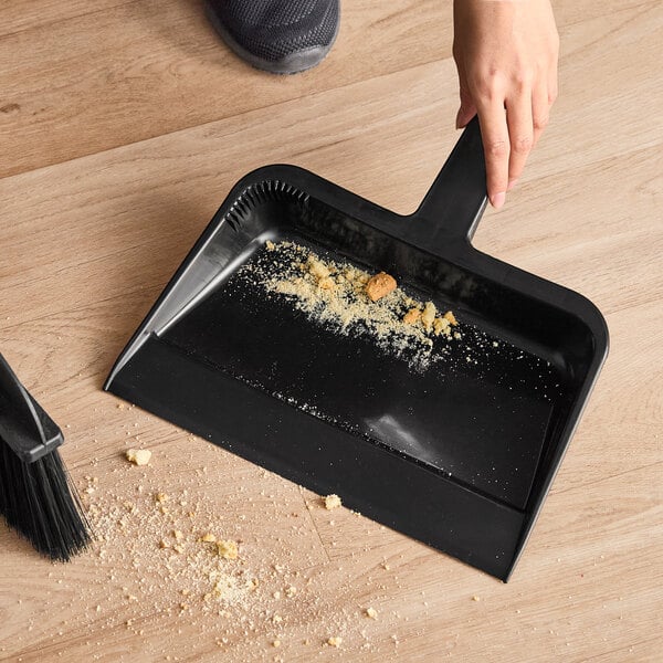 A black plastic utility dust pan being used to collect crumbs from a wooden floor.