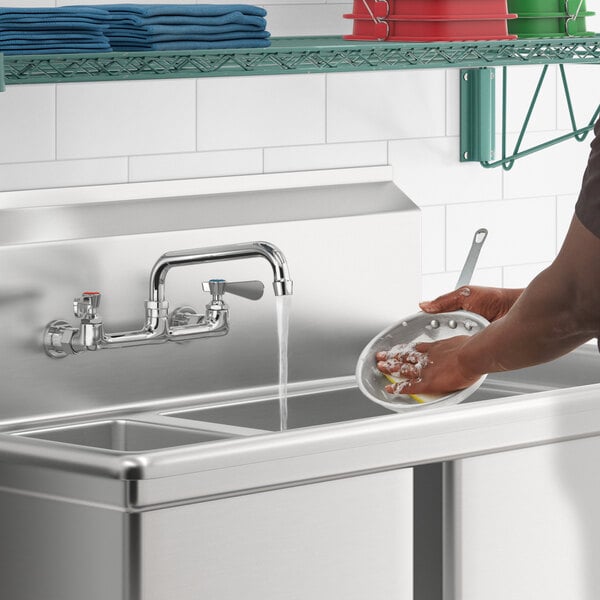 A person using a Regency wall mount faucet to wash dishes in a stainless steel sink.