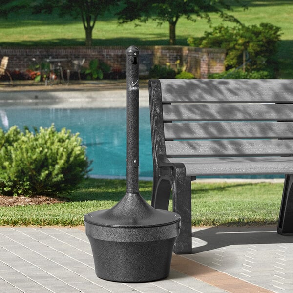 A black Rubbermaid Aladdin cigarette receptacle on a metal pole by a bench in a park.