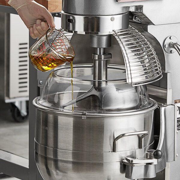 A person's gloved hand pouring brown liquid from a measuring cup into an Avantco mixer with a splash cover.