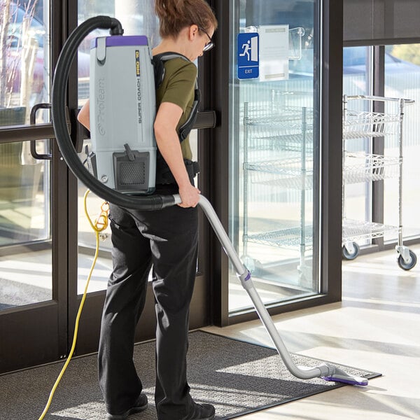 A woman using a ProTeam backpack vacuum with a white rectangular attachment to vacuum a carpet in a corporate office.