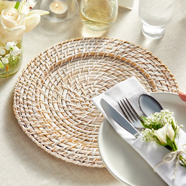 A white plate on a blond rattan charger with silverware and a napkin.