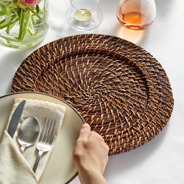 A hand holding an Acopa Rattan charger plate with a napkin and a fork.