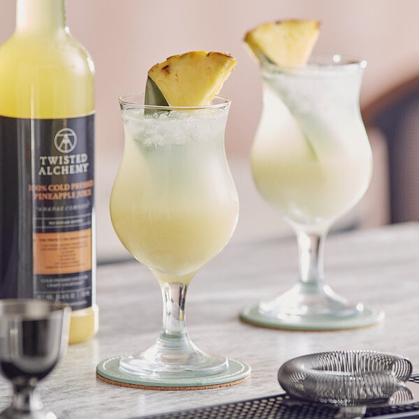 A bottle of Twisted Alchemy Cold-Pressed Pineapple Juice on a table with two glasses of yellow liquid and pineapple slices.