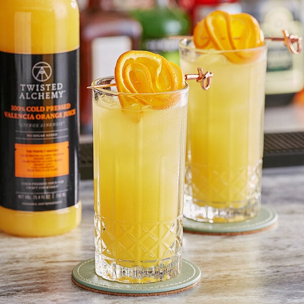 Two glasses of Twisted Alchemy Valencia orange juice with orange slices on top.