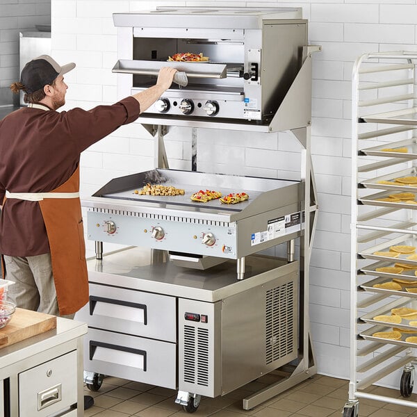 A man in a brown apron using a Regency Salamander broiler stand to cook food.