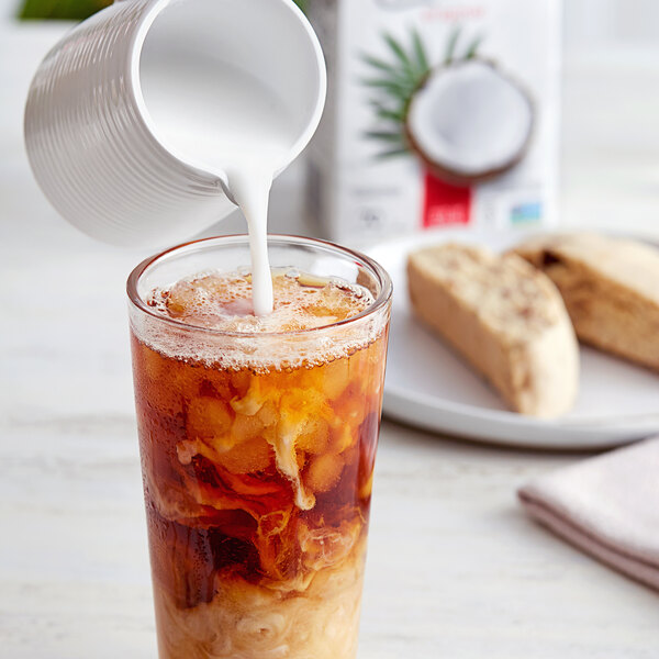 A white jug pouring So Delicious Organic Coconut Milk into a glass of brown liquid.