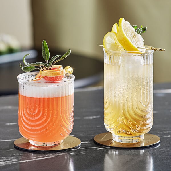 Two Acopa Zelda highball glasses with orange and yellow cocktails and fruit garnishes on a table.