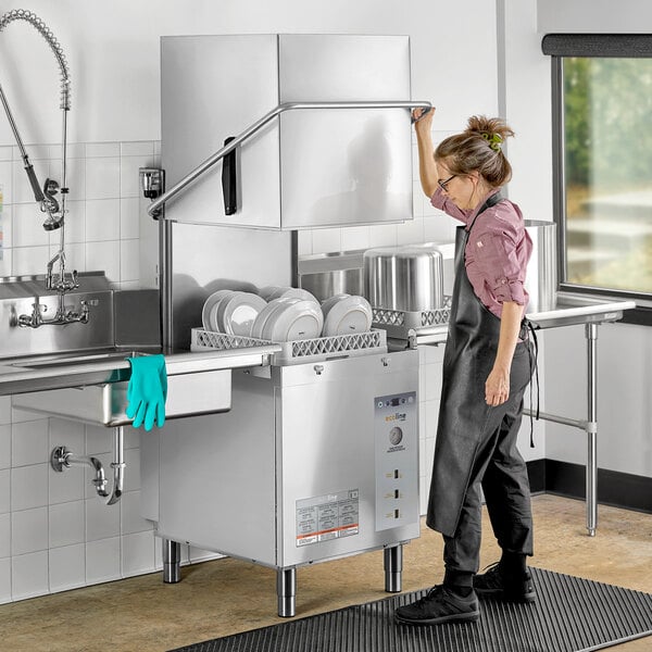 A woman in overalls stands in a professional kitchen next to a Ecoline by Hobart door-type dishwasher.
