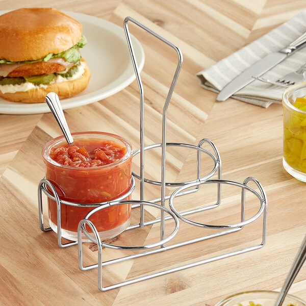 A metal wire condiment holder with a jar of sauce next to a burger.