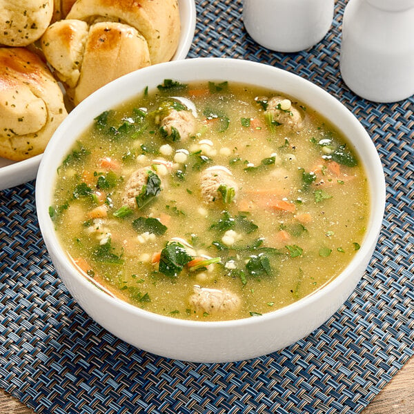 A bowl of Chef Francisco Italian-Style Wedding Soup with meatballs and vegetables next to bread rolls.