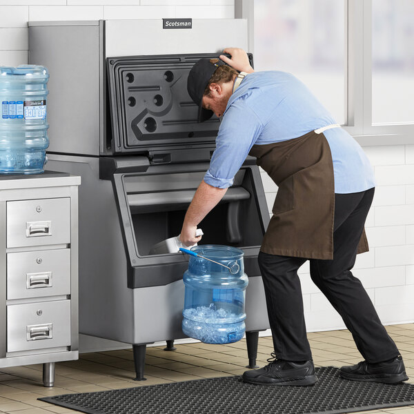 A man in an apron using a water hose to put water into a black square plastic container on a counter.