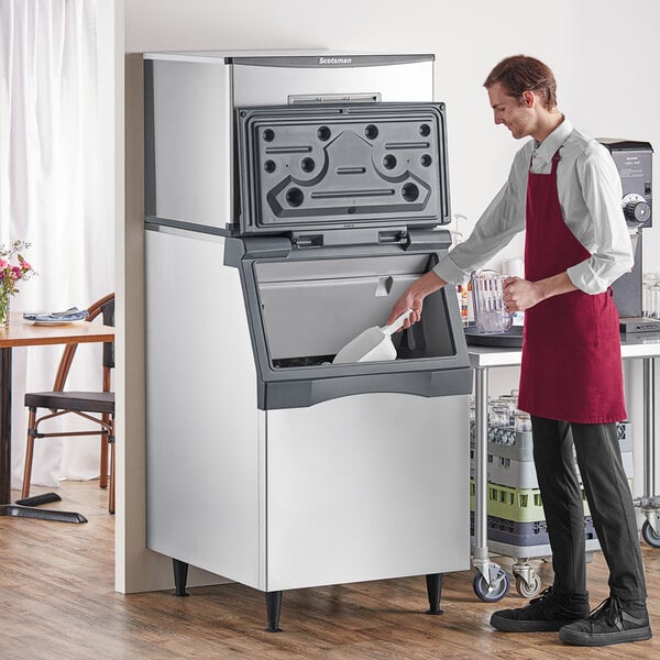 A man in a red apron using a white scoop to get ice from a large white Scotsman ice machine.