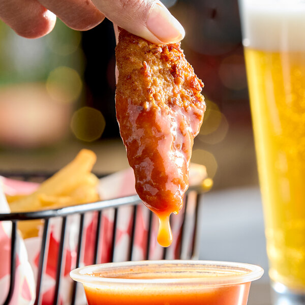A piece of fried chicken being dipped into a cup of orange habanero hot sauce.