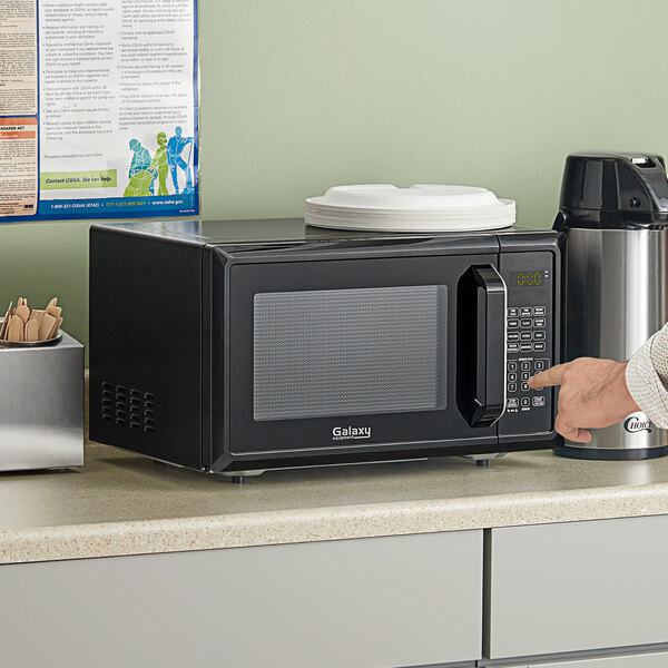 A person pointing at a black Galaxy office series microwave on a counter.