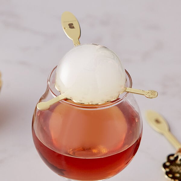 A hand holding a Flavour Blaster Gold Bubble Spoon over a glass of liquid on a table.