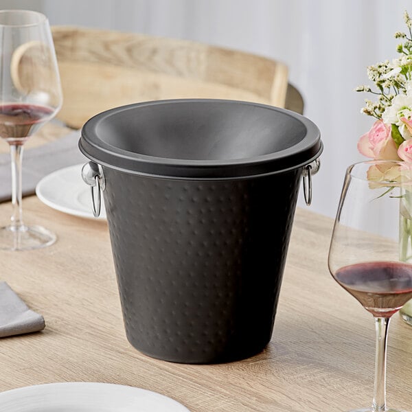 An Acopa black hammered stainless steel wine tasting spittoon on a table.