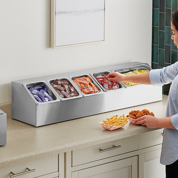 A woman standing at a counter with food in containers, reaching for a stainless steel pan organizer with condiment pans inside.