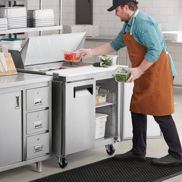 An apron-wearing man using gloves to put food into an Avantco 2 door sandwich prep refrigerator.