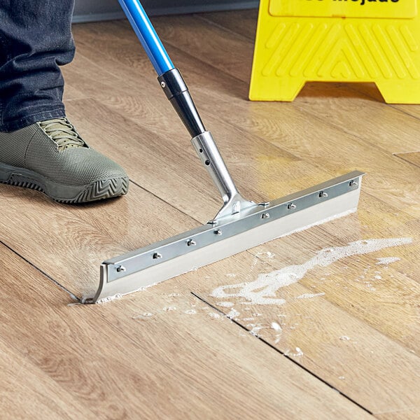 A 24-inch rubber floor squeegee with a metal frame and a 60-inch tapered wood handle being used to clean liquid from a wooden floor.