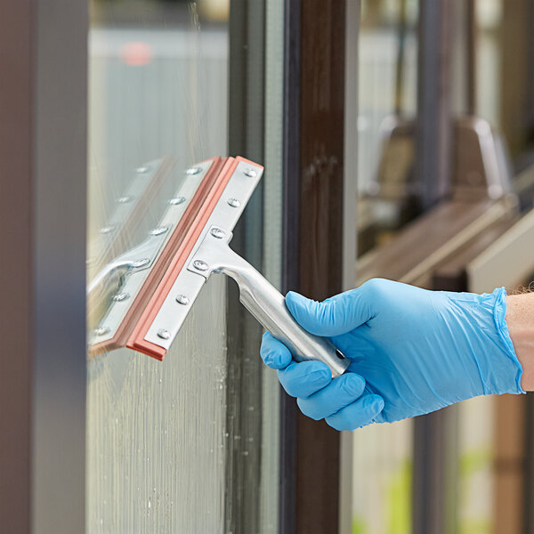 A hand in blue gloves using a Lavex squeegee with a metal blade to clean a window.