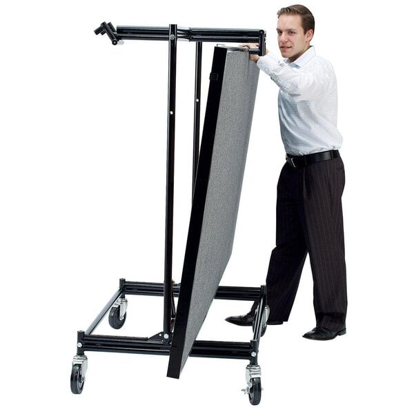 A man in a white shirt and black pants pushing a large black National Public Seating stage dolly.