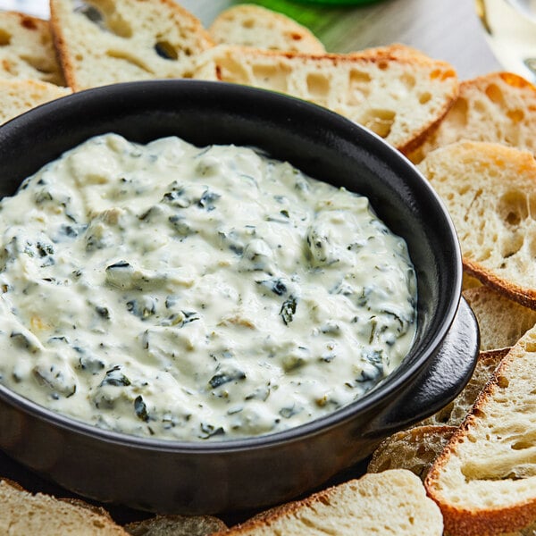 A bowl of Kettle Collection Parmesan Spinach Artichoke Dip on a plate with bread and crackers.