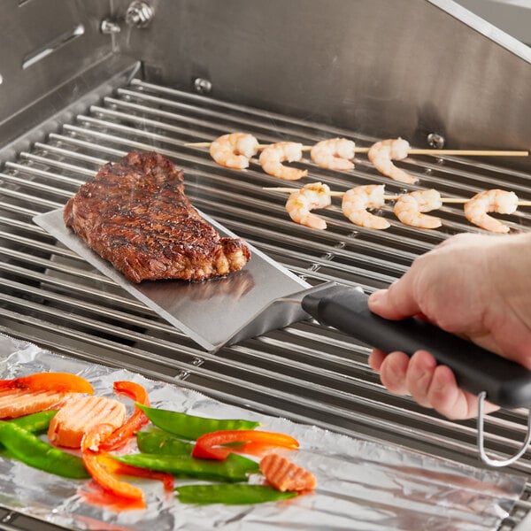 A hand holding a Backyard Pro High Heat Solid Turner with a black handle over meat on a grill.