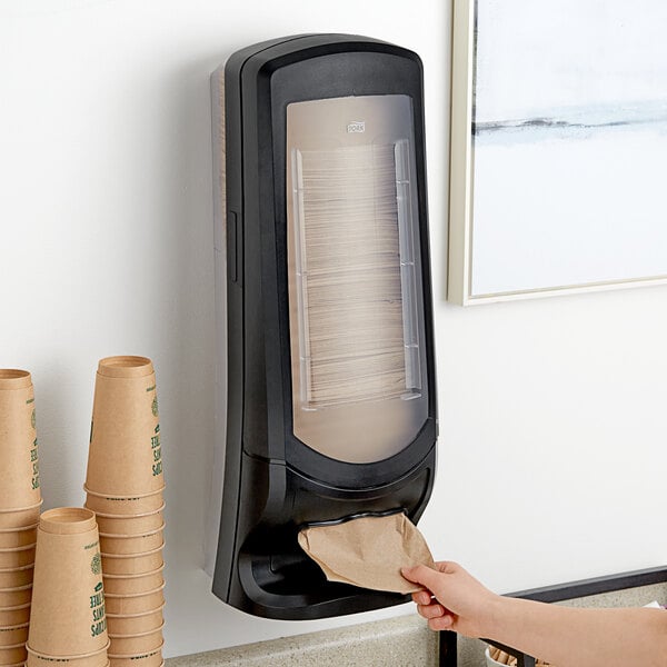 A person putting a brown paper bag into a black Tork Xpressnap standing wall mount dispenser.