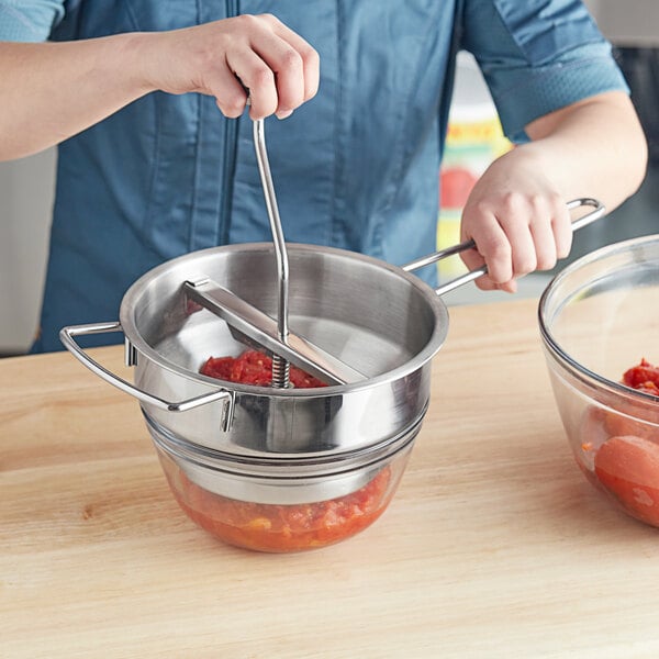 A person uses the stainless steel rotary food mill to make food in a metal bowl.