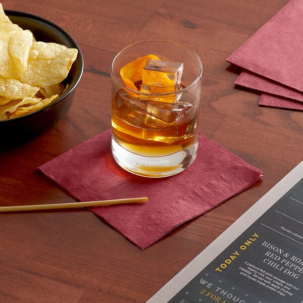 A bowl of chips and a glass of liquid on a table with a Choice Burgundy beverage napkin.