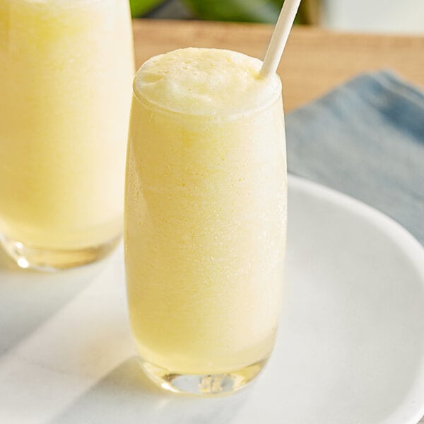 Two glasses of yellow smoothies with straws on a white plate.
