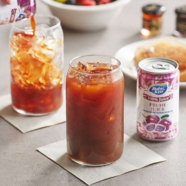 A glass of Ruby Kist prune juice with ice on a table with a bowl of fruit.