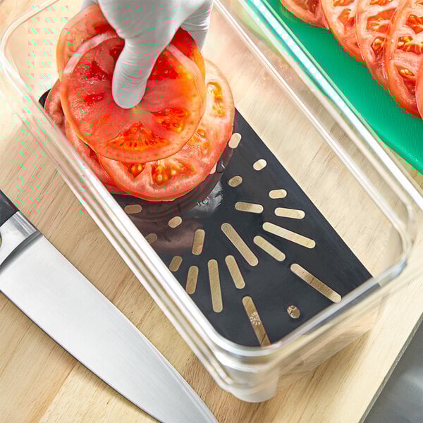 A person cutting a tomato into a black Cambro polycarbonate container over a table.