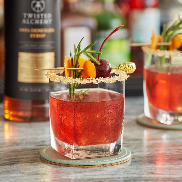 A bottle of Twisted Alchemy Demerara Syrup on a table with two cocktails garnished with fruit and rosemary.