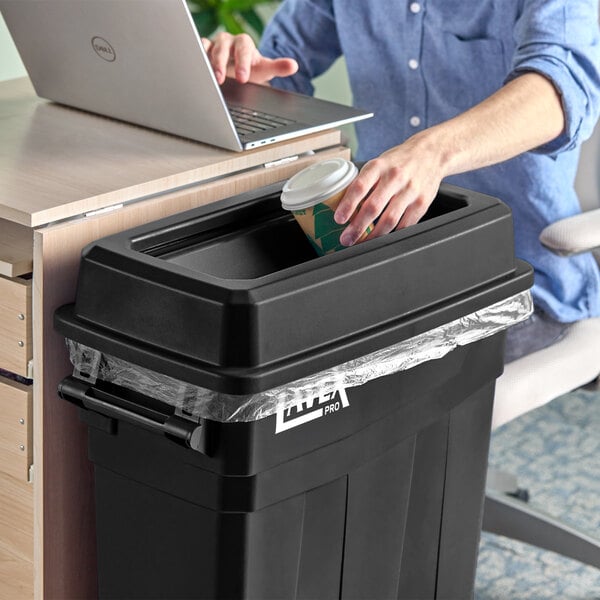 A person using a Lavex Pro black slim rectangular trash can to put a coffee cup in.