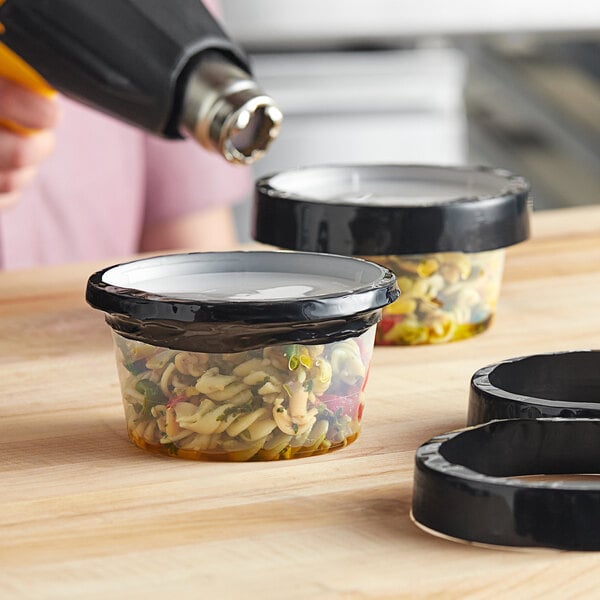 A person using a blow dryer to shrink a black band on a container of food.