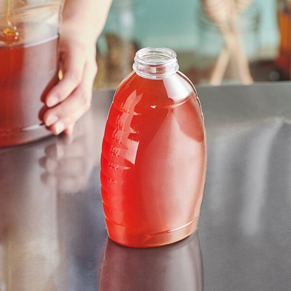 A hand pouring honey from a Classic Queenline PET Honey Bottle into a glass of brown liquid on a table.