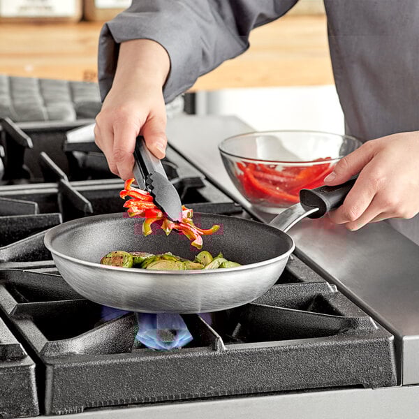 A person cooking vegetables in a Vollrath Wear-Ever aluminum non-stick fry pan with a black silicone handle.