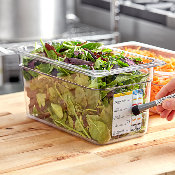 A hand using a white marker to write on a Carlisle clear food pan filled with salad.