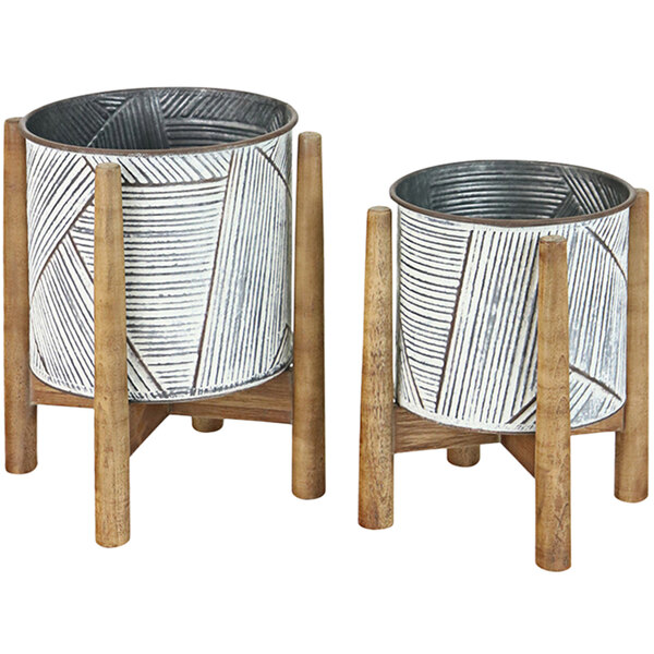 A white metal planter with a gray design on a wooden stand next to a white metal planter with a white design.