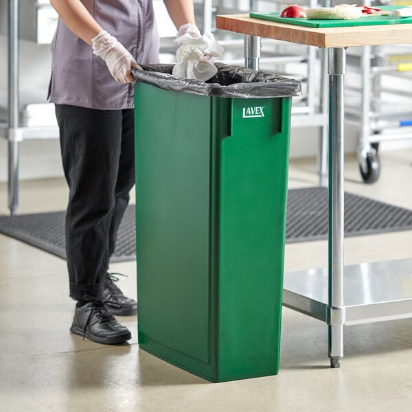 A woman standing next to a Lavex dark green rectangular trash can on a counter.