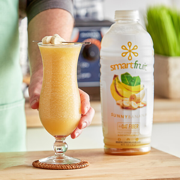 A person's hand holding a glass of yellow Smartfruit Sunny Banana beverage with a banana slice on the rim.